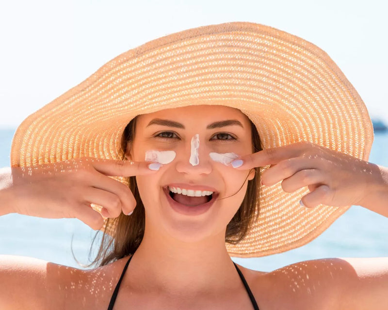 Smiling woman in a wide-brim sun hat applying sunscreen to her nose at the beach