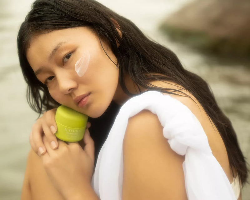 Young Asian woman holding a Kayura Barrier repair cream jar with a white towel draped over her shoulder outdoors