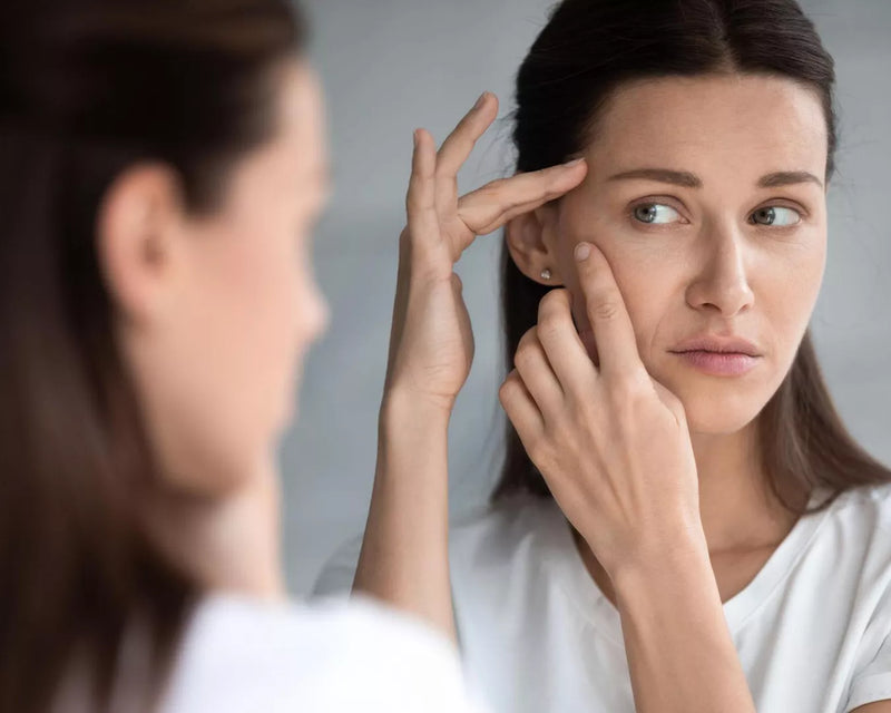 Woman in a white shirt examining her face closely in a mirror with fingertips pressed to her cheek
