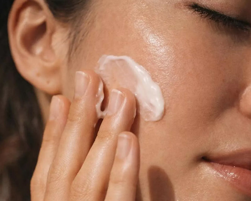 Close-up of a woman applying white moisturiser cream to her cheek with fingertips