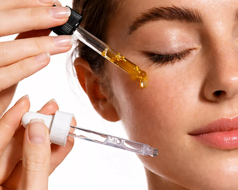 Close-up of a glass dropper applying golden serum drops under a woman's eye