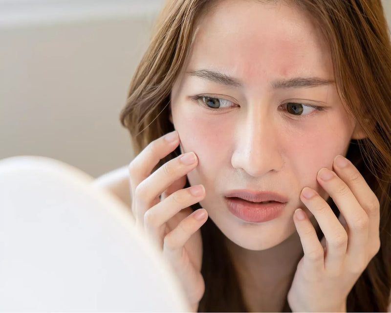 Asian woman touching her cheeks while examining her skin closely in a mirror