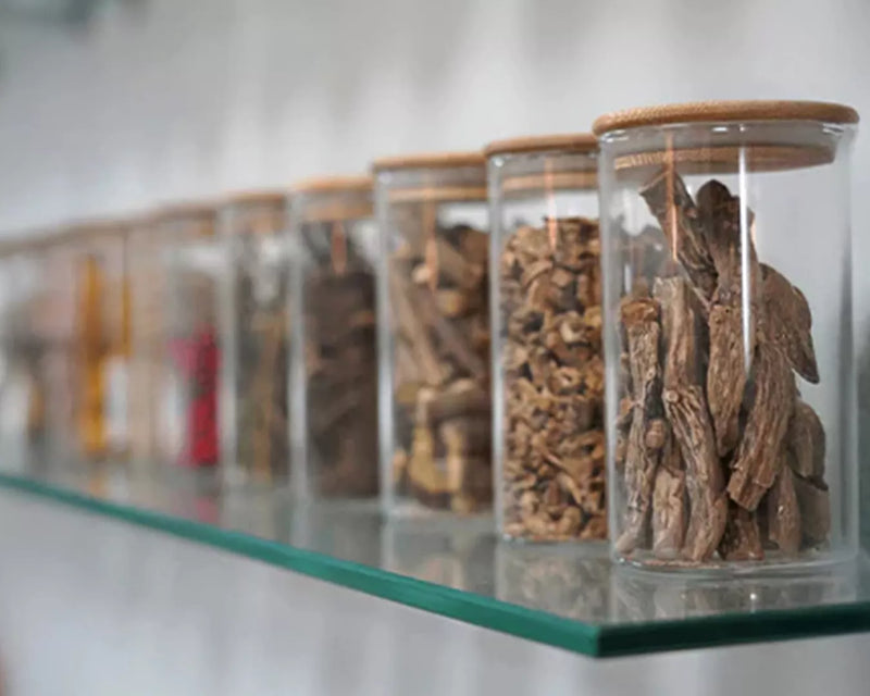 Row of glass storage jars filled with dried roots and botanical herbs on a white shelf