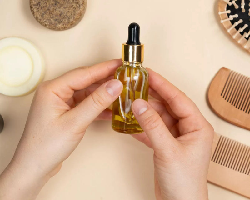 Hands holding a small amber glass dropper oil bottle beside a wooden hair comb on a beige surface
